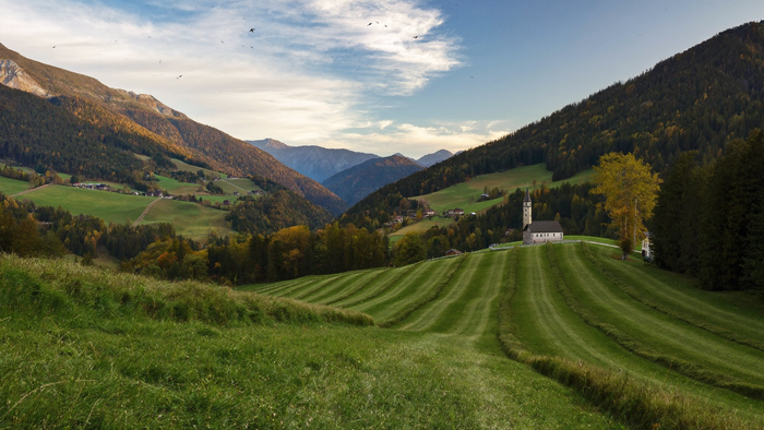 Herbstliche Bergwiese in einem Alpental mit gemähten Streifen, dahinter Kirche und Wald im Abendlicht – KI-Illustration, nicht der Originalort des Monuments
