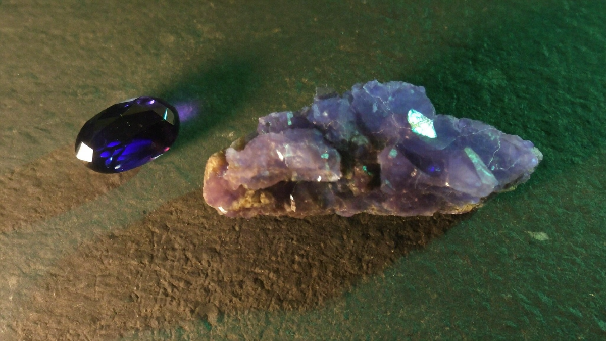 Iolith: violettblauer facettierter Edelstein neben einem violetten Rohstein auf dunklem Schiefer im Kerzenlicht