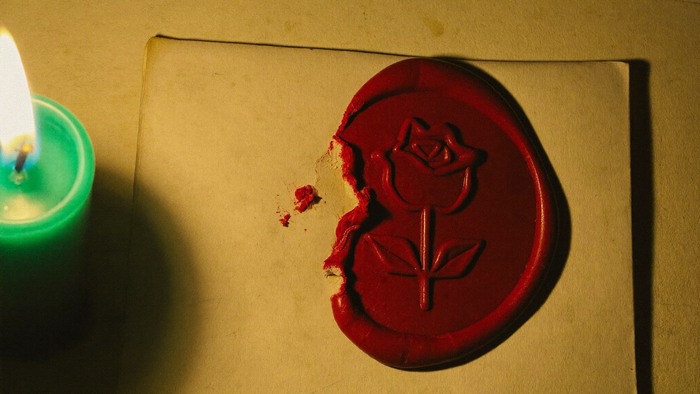 Rotes Wachssiegel mit Rosenprägung, am Rand aufgebrochen, auf hellem Papier neben einer brennenden Kerze