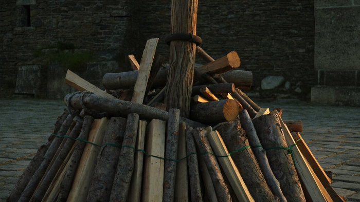 Unentzündeter Scheiterhaufen: dicker Holzpfahl mit dunklem Ring, rundum gestapelte Scheite und Reisig auf Kopfsteinpflaster