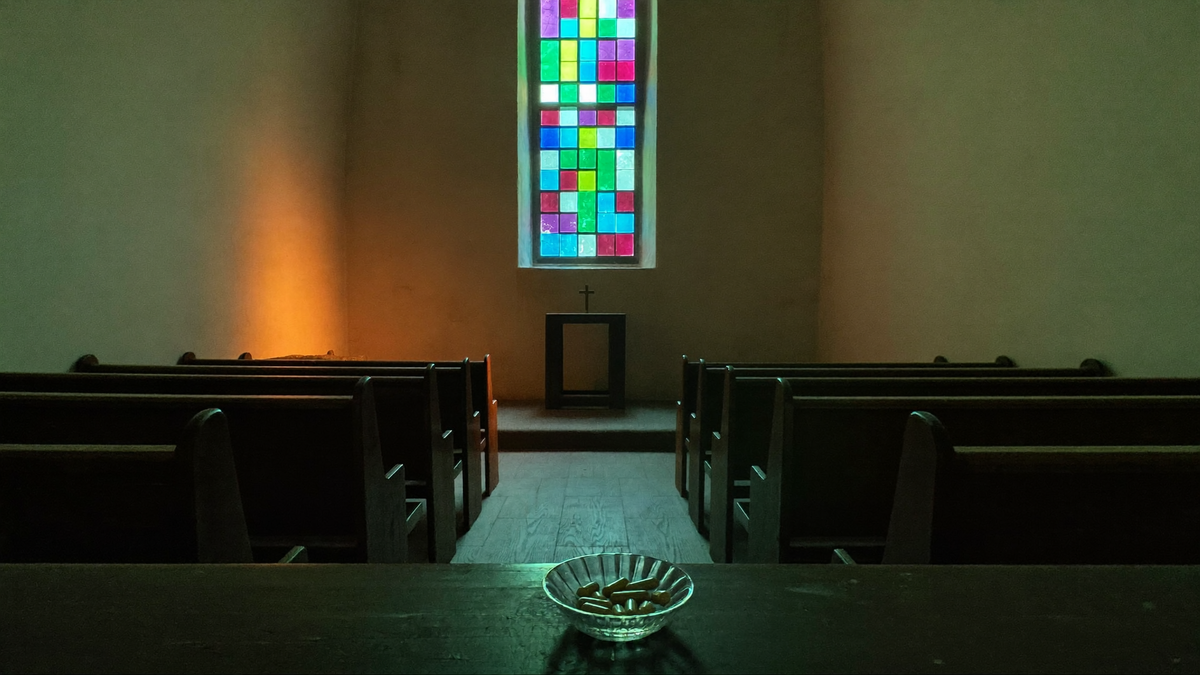 Leere kleine Kapelle mit dunklen Holzbänken, schlichtem Altar mit Kreuz und buntem Glasfenster, seitlich warmes Licht; vorne auf der Bank eine Glasschale mit Kapseln