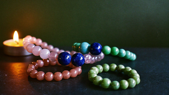Drei Edelstein-Armbänder für Kinder aus Rosenquarz, Aventurin und Lapislazuli auf dunklem Stein, daneben eine brennende Kerze