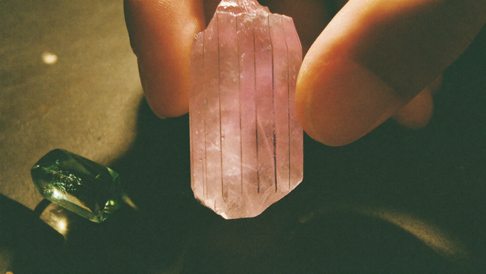 Fingerspitzen halten einen blassrosa Kunzit-Kristall mit feinen Längsstreifen gegen das Licht, darunter ein grüner Hiddenit im Kerzenlicht