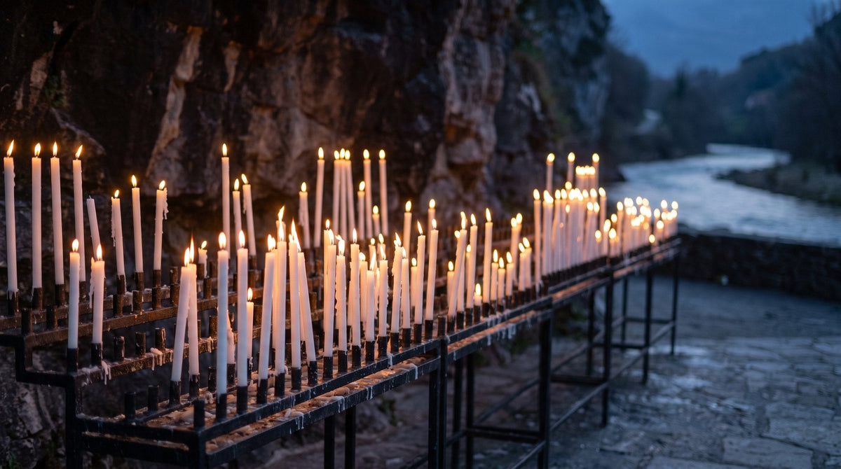 Brennende weiße Opferkerzen auf eisernen Kerzenständern vor einer Felswand, dahinter ein Fluss in der Abenddämmerung – KI-Illustration, nicht die Grotte von Lourdes