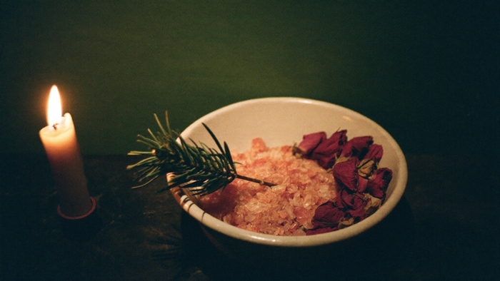 Keramikschale mit grobem Badesalz, getrockneten Rosenblüten und einem Fichtenzweig neben einer brennenden Kerze