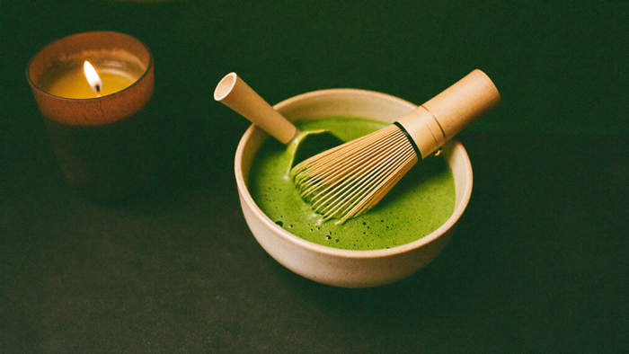 Schale mit aufgeschlagenem Matcha, Bambusbesen und Bambusgriff auf dunkler Fläche im warmen Kerzenlicht