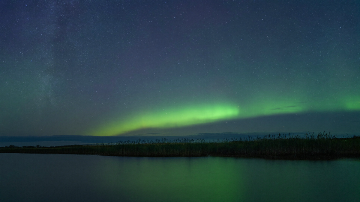 Nachtlandschaft an einer flachen norddeutschen Küste, über dem Nordhorizont ein niedriger grüner Polarlichtbogen, darüber Sternenhimmel