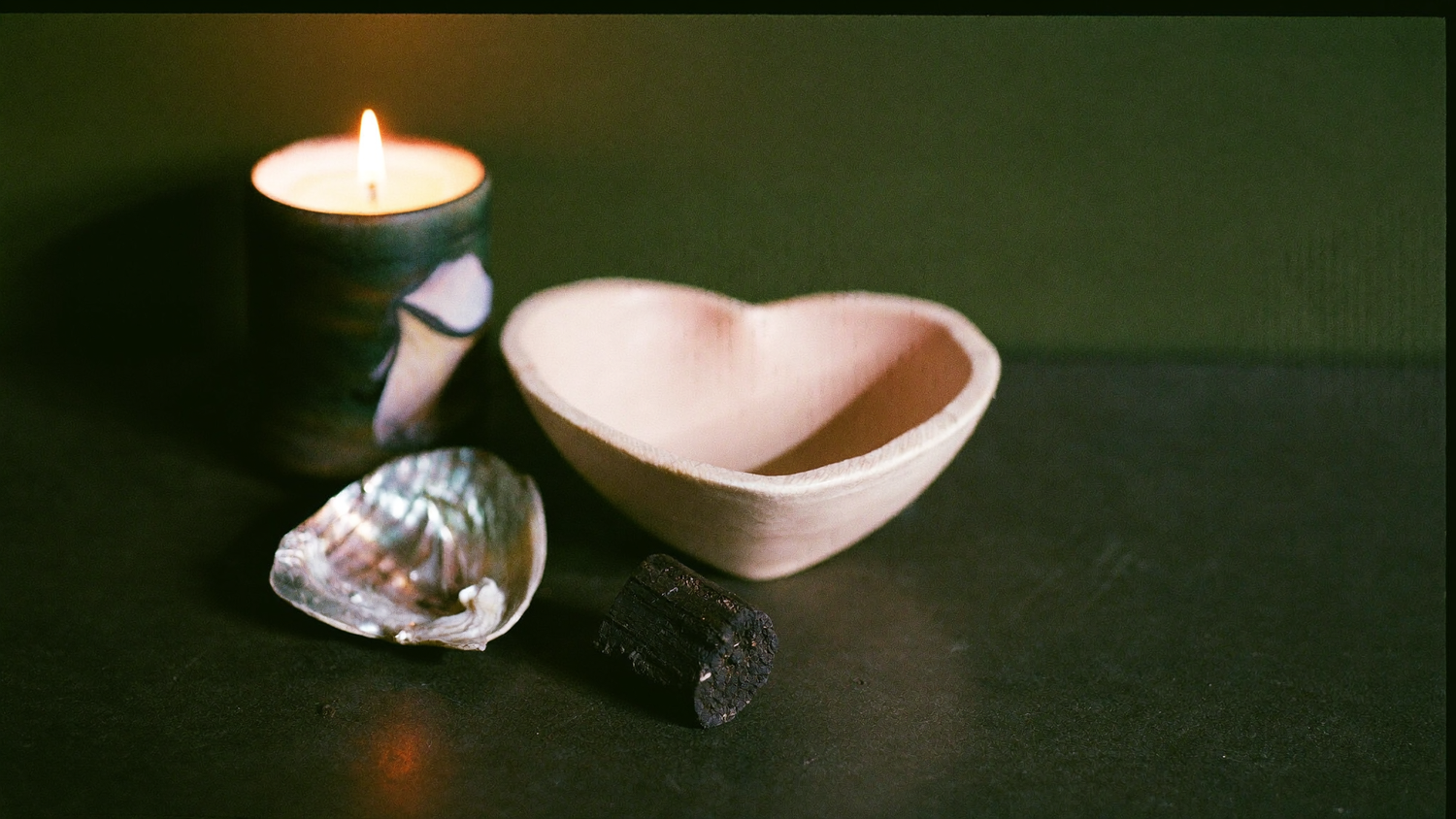 Herzförmige Keramikschale, Abalone-Muschel und Palo Santo Holz auf dunklem Stein neben einer brennenden Kerze