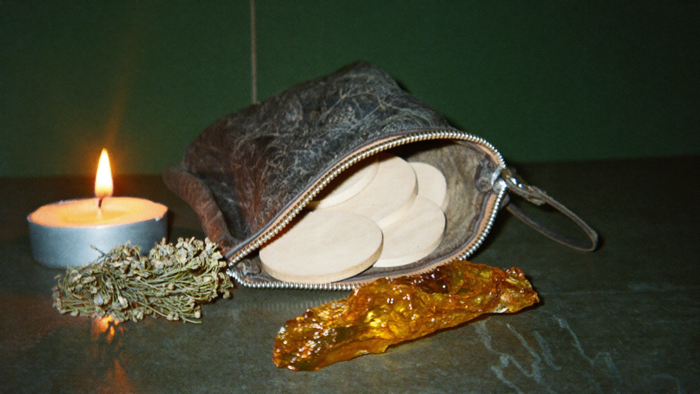 Geöffneter Lederbeutel mit hellen Holzplättchen, daneben getrocknetes Heidekraut, ein Stück roher Bernstein und eine brennende Kerze
