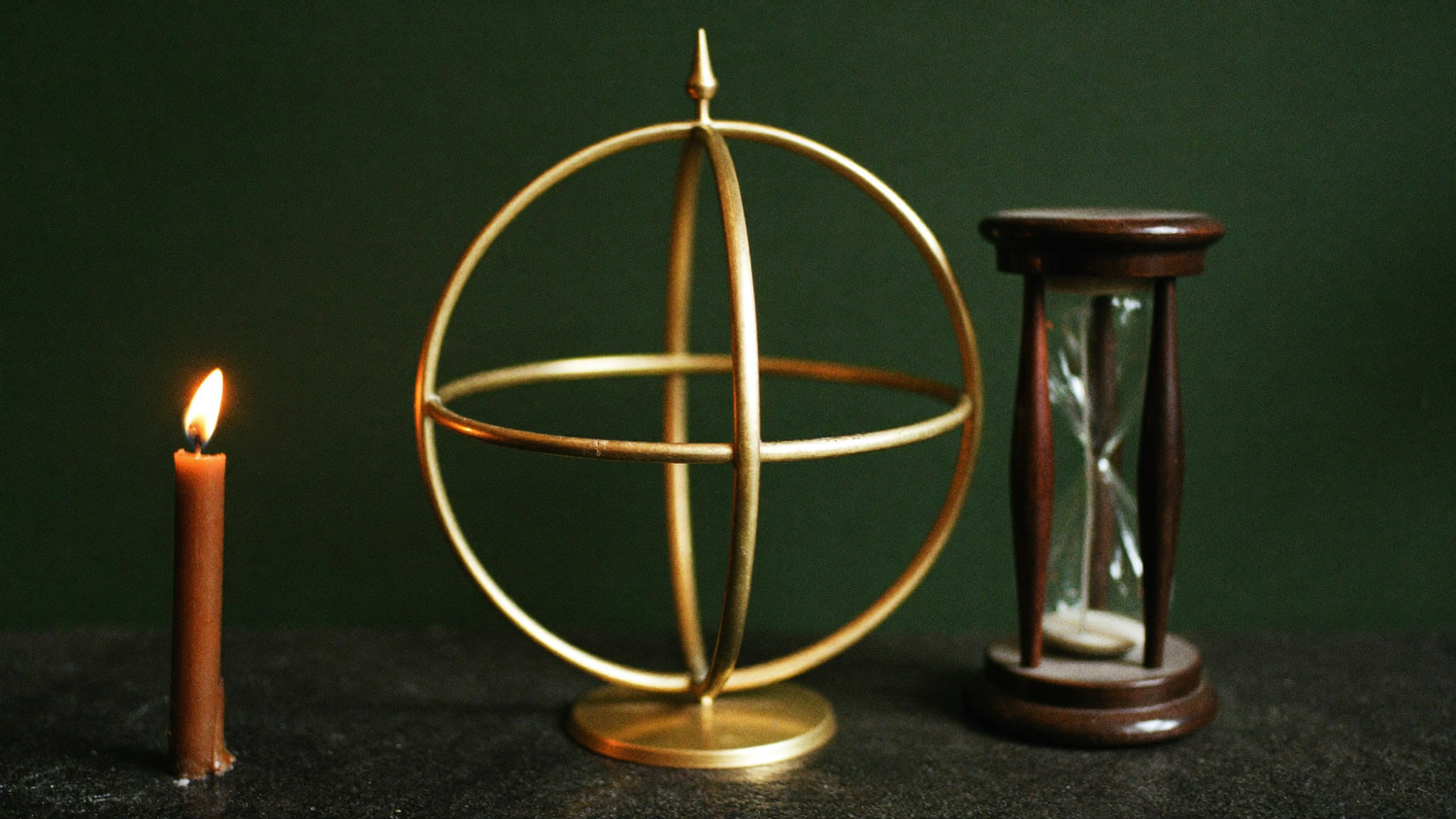 Eine schlichte Armillarsphäre aus Messing steht neben einer kleinen Sanduhr auf dunklem Stein, links brennt eine Kerze vor einer dunkelgrünen Wand.