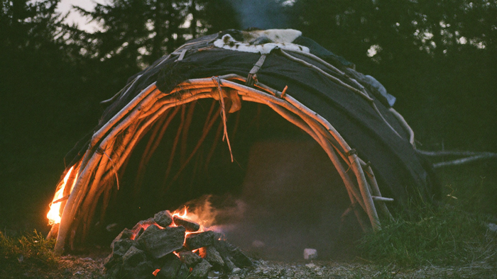Schwitzhütte aus gebogenen Weidenruten mit dunklen Decken und Fellen, davor ein Haufen glühender Steine im Feuer in der Dämmerung