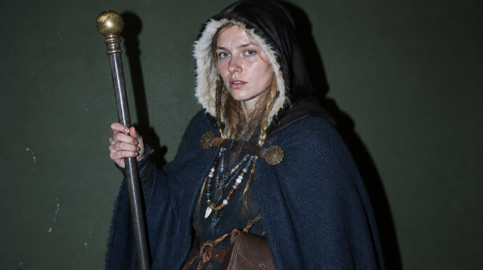 Seiðr: junge Frau als Völva mit Eisenstab und Messingknauf, blauem Wollmantel, schwarzer Lammfellhaube mit weißem Futter, Glasperlenketten und Lederbeutel