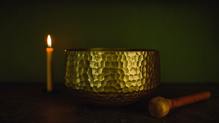 Gehämmerte Bronze-Klangschale mit Holzschlägel neben einer brennenden Kerze vor dunkelgrüner Wand