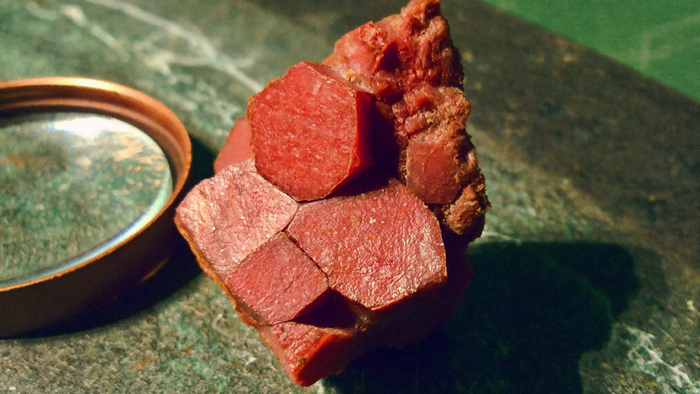 Roter Spinell-Rohkristall mit scharfen Flächen neben einer Juwelierlupe auf dunklem Stein im Kerzenlicht — Nahaufnahme