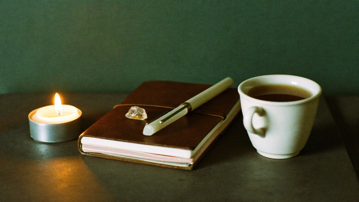 Geschlossenes Ledertagebuch mit Stift, kleinem Bergkristall und einer Tasse Tee neben einer brennenden Kerze vor dunkelgrüner Wand