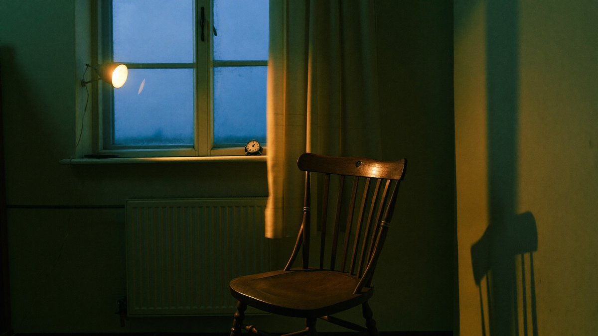 Ein leeres Zimmer zur blauen Stunde: ein Holzstuhl am hohen Fenster, eine kleine Uhr auf dem Fensterbrett, eine Lampe wirft warmes Licht