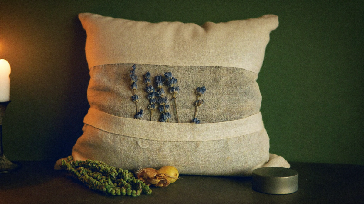 Leinenkissen mit getrocknetem Lavendel, Hopfenzapfen und Kräutern bei Kerzenlicht vor dunkelgrüner Wand — Titelbild zum Traumkissen