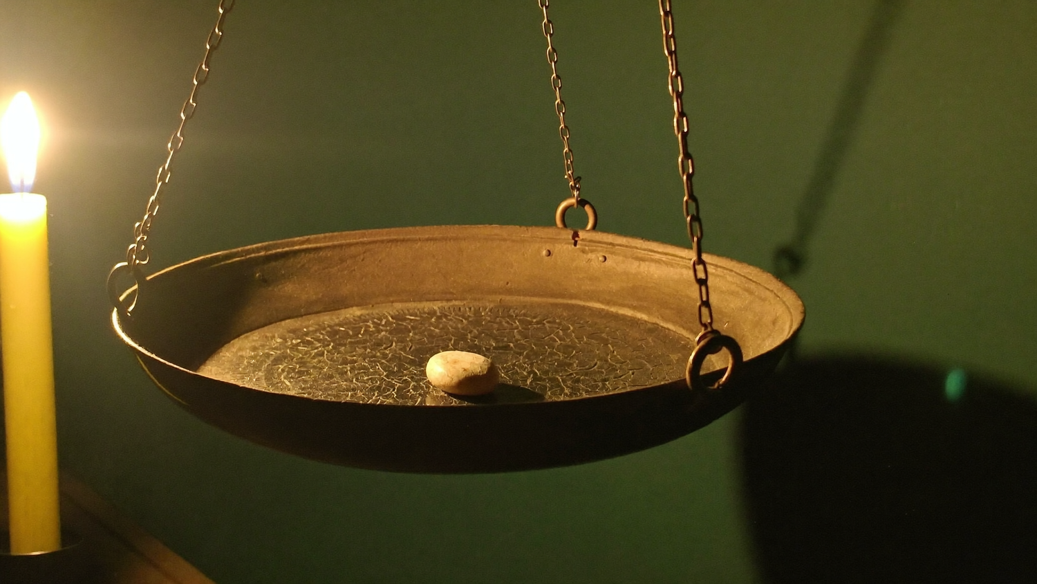Messingschale einer alten Balkenwaage an Ketten mit einem einzelnen hellen Stein, links eine brennende Kerze vor dunkelgrüner Wand