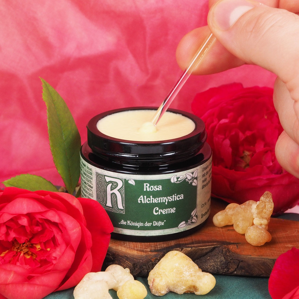 Geöffneter Tiegel Rosa Alchemystica Creme, mit einem Spatel wird helle Creme entnommen, daneben Rosen und Weihrauch