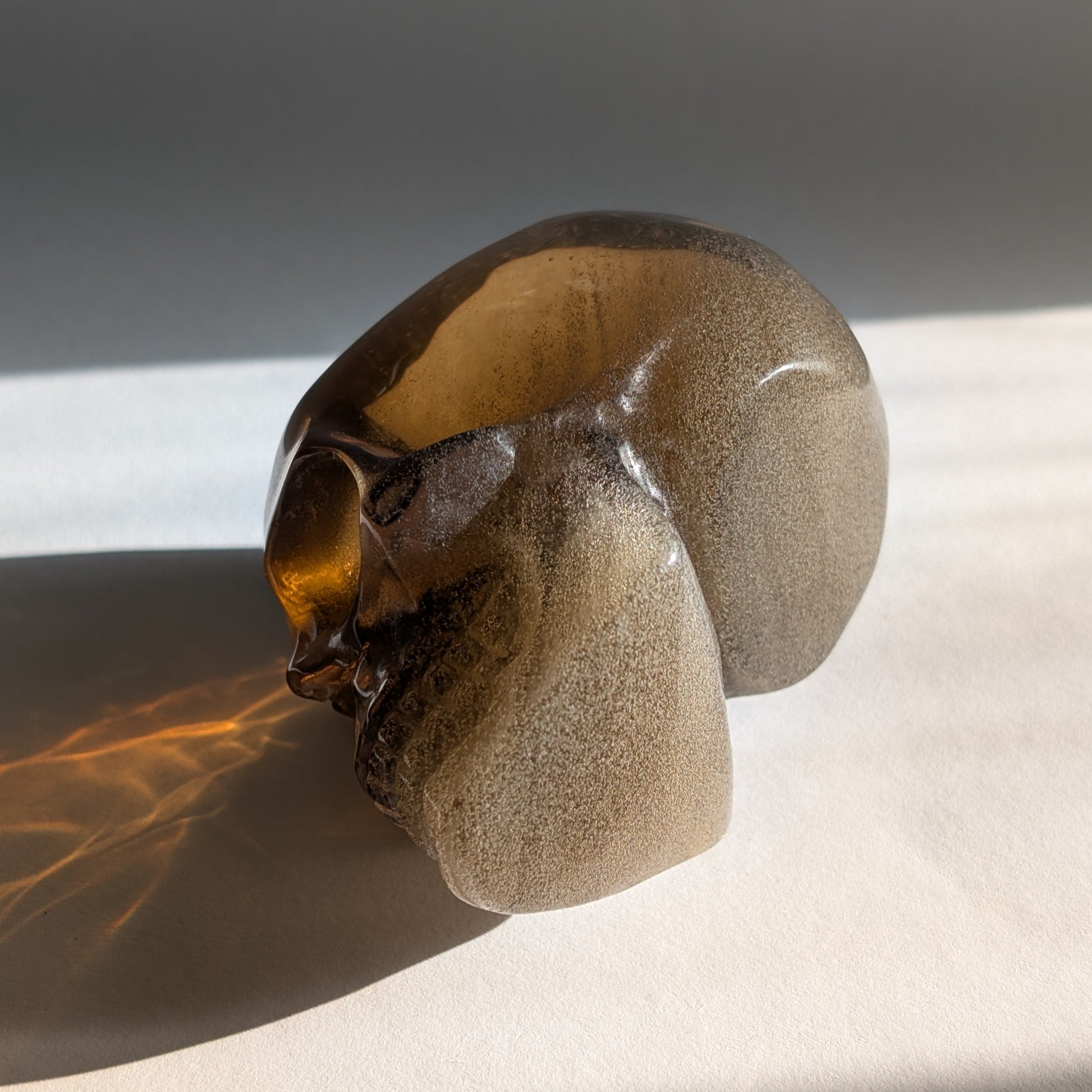 Ein 258g schwerer, synthetischer Rauchquarz-Schädel 'Auric' mit unzähligen goldenen Partikeln, die wie Sternenstaub im Licht funkeln.