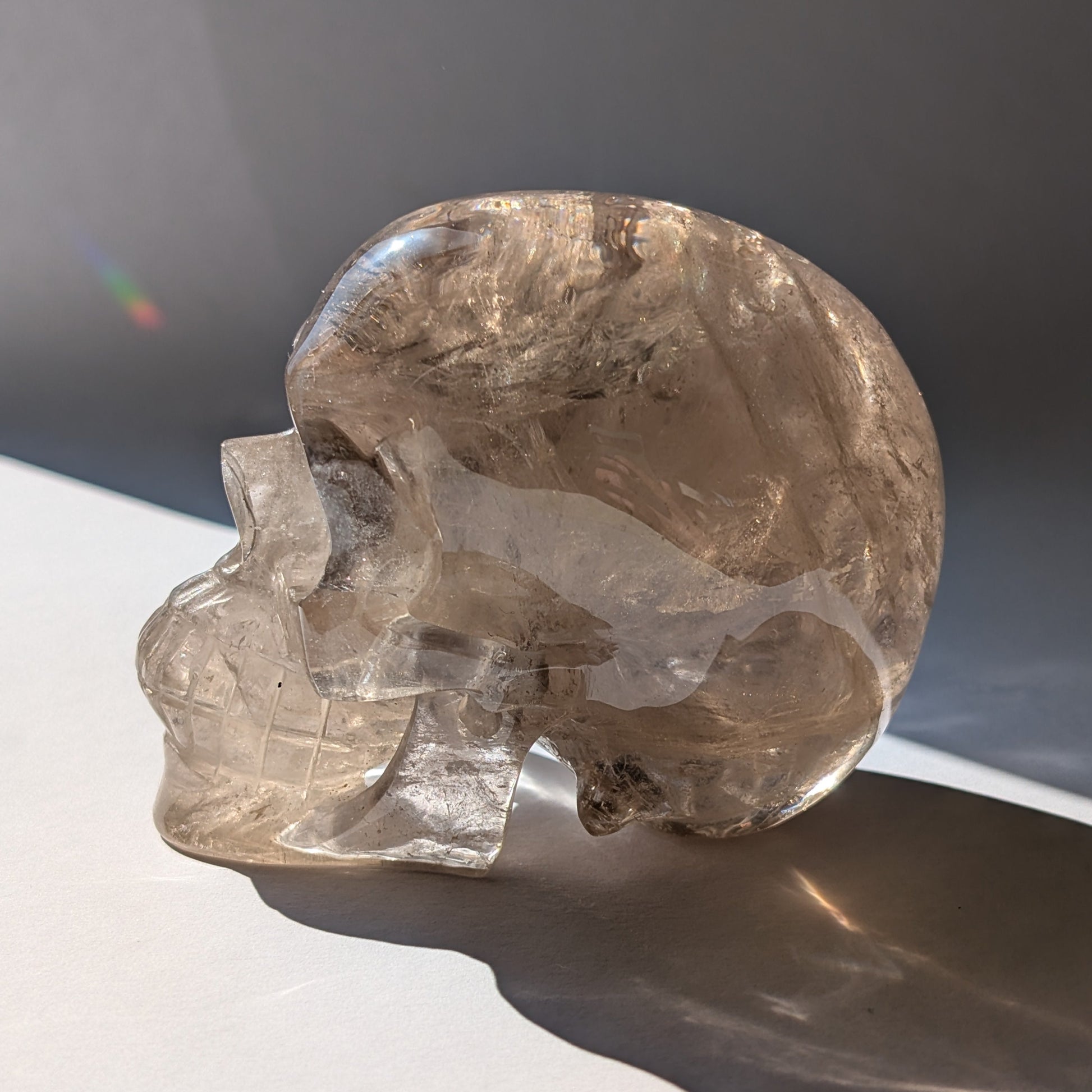 Ein 594g schwerer, klarer Rauchquarz-Schädel 'Der Wächter der Schatten' mit leuchtenden Regenbogen-Einschlüssen.