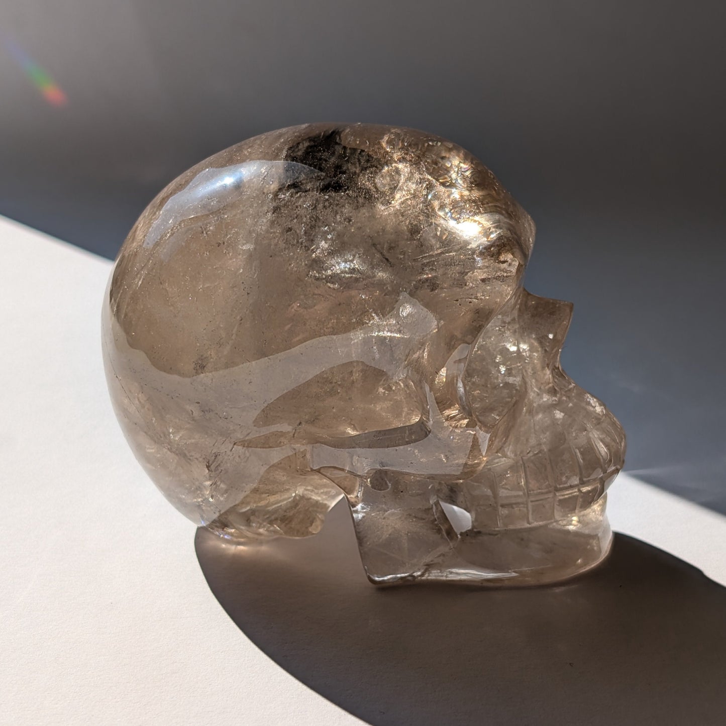 Ein 594g schwerer, klarer Rauchquarz-Schädel 'Der Wächter der Schatten' mit leuchtenden Regenbogen-Einschlüssen.
