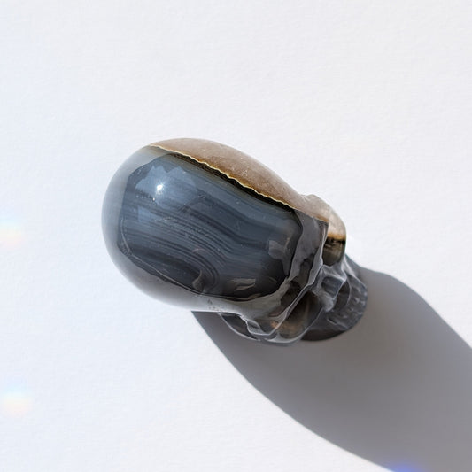 Ein 86g schwerer Kristallschädel 'Der Wächter der zwei Welten' aus Gebändertem Achat, dessen eine Hälfte eine offene, funkelnde Druzy-Geode ist.