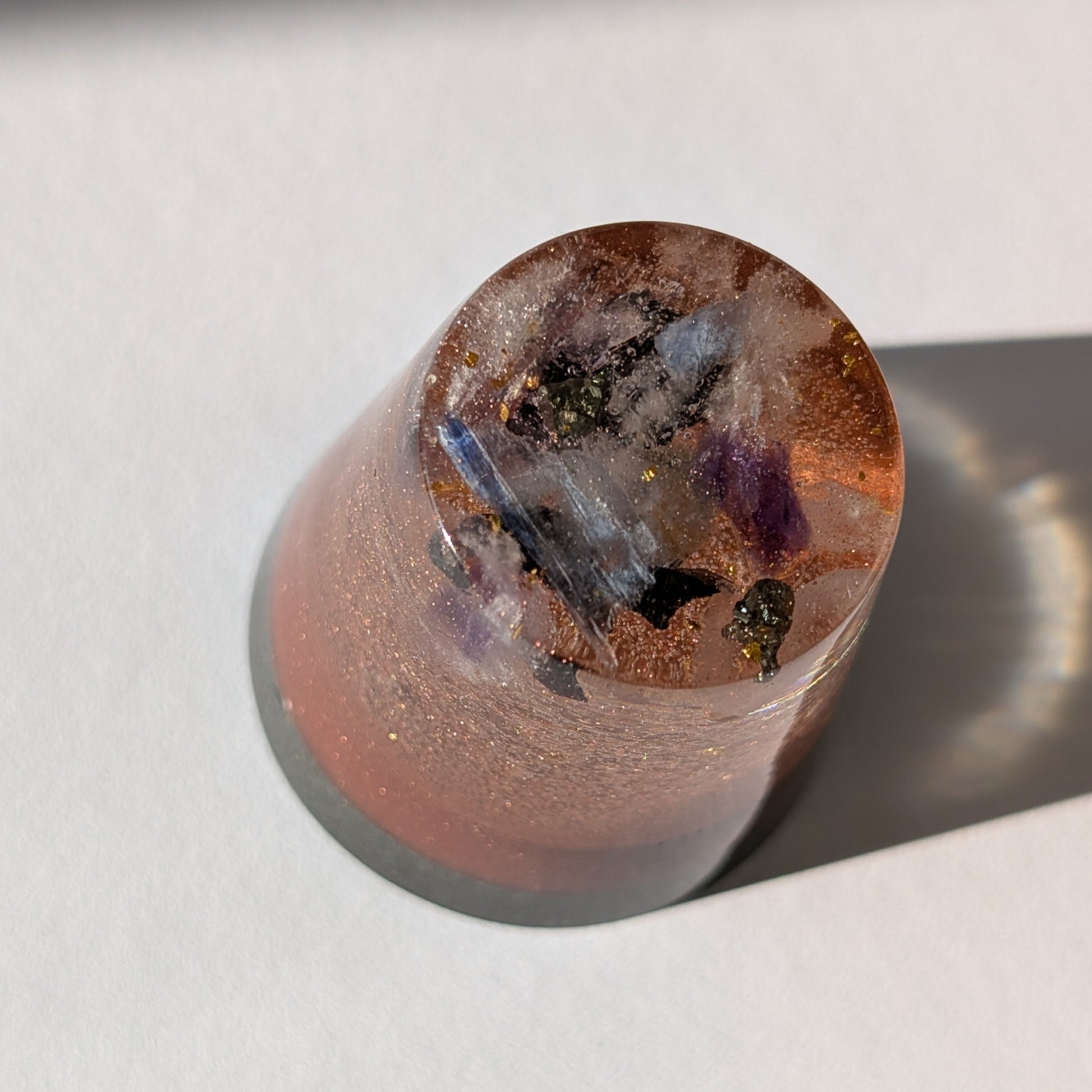 Ein kleiner, kupferfarbener Orgonit Kegel 'Alchemisten-Funke' von Einklang Manufaktur, der im Licht funkelt und Kristalle im Inneren zeigt.