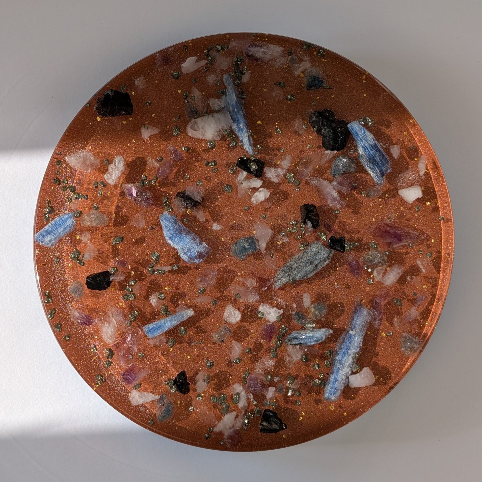 Eine große, runde Orgonit Energetisierungsplatte (17cm) 'Quelle der Kraft' mit sichtbaren Kristallen wie Kyanit und Pyrit in einer rötlichen Matrix.