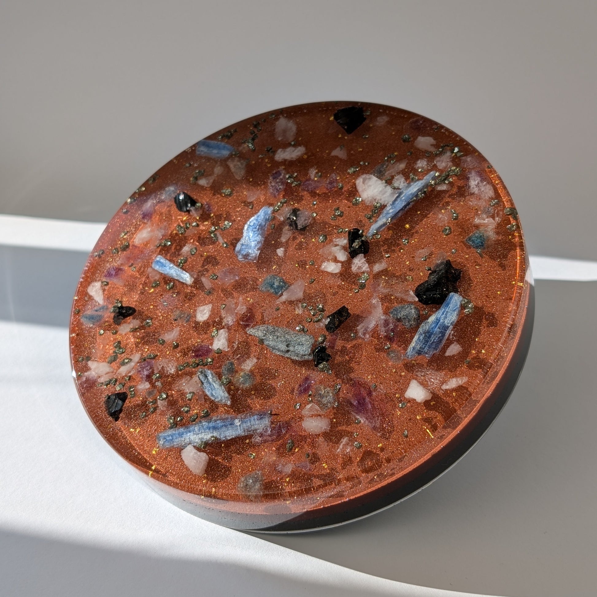 Eine große, runde Orgonit Energetisierungsplatte (17cm) 'Quelle der Kraft' mit sichtbaren Kristallen wie Kyanit und Pyrit in einer rötlichen Matrix.