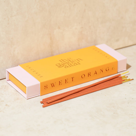 Eine orangefarbene Box von The Golden Altar mit 30 orangefarbenen Süße Orange Räucherstäbchen, die daneben liegen.