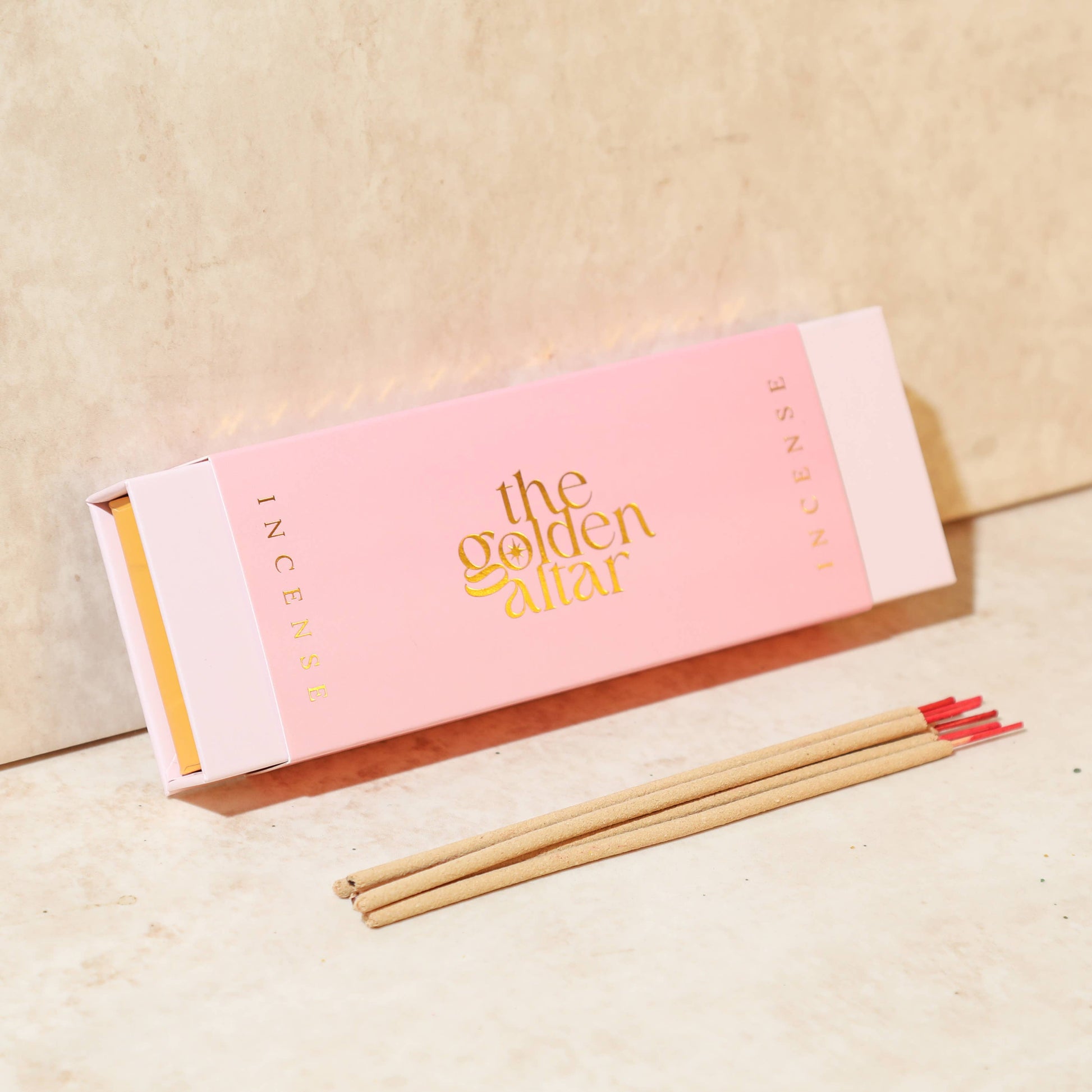Eine rosa-orangefarbene Box von The Golden Altar mit 30 Rose & Vetiver Räucherstäbchen, die daneben liegen.