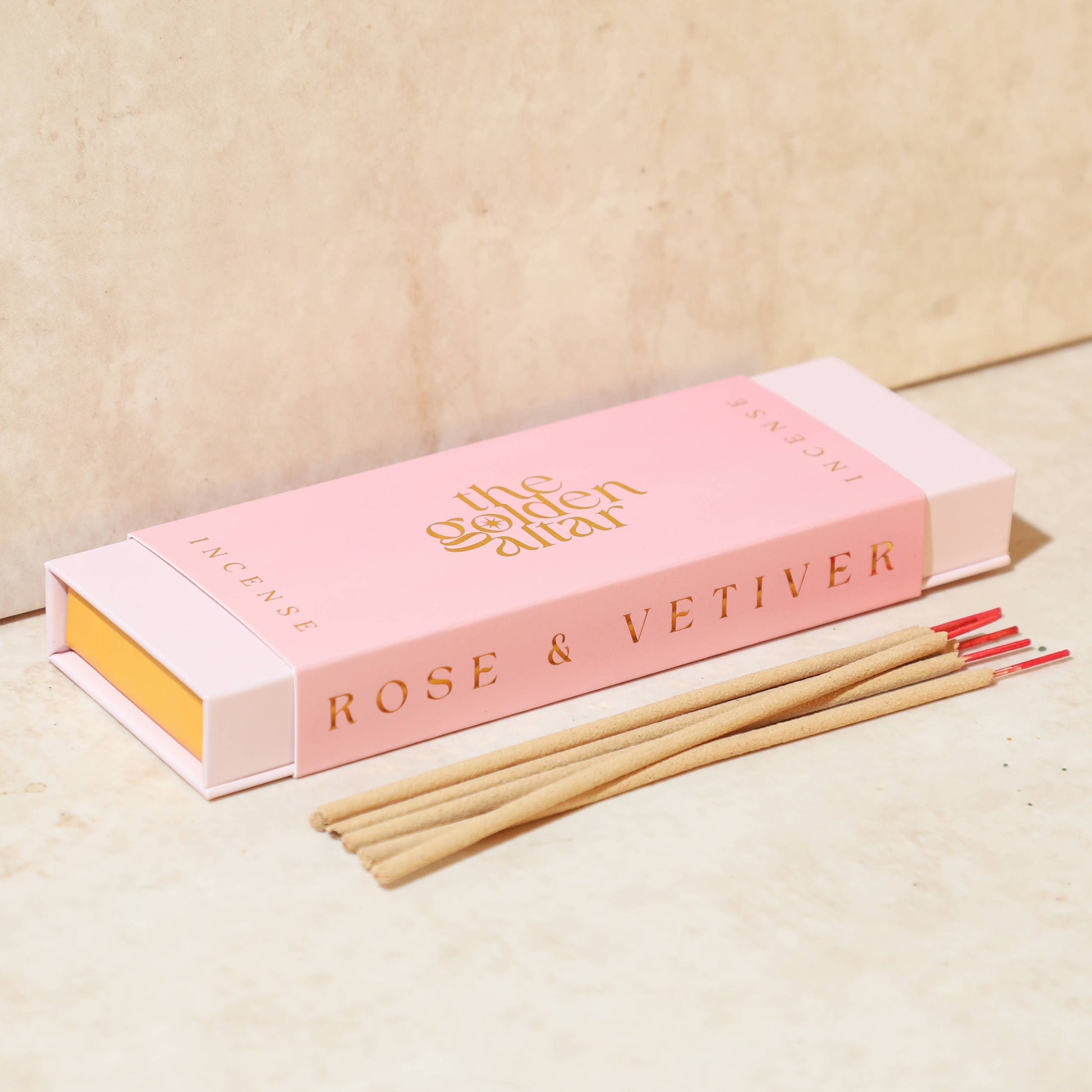 Eine rosa-orangefarbene Box von The Golden Altar mit 30 Rose & Vetiver Räucherstäbchen, die daneben liegen.