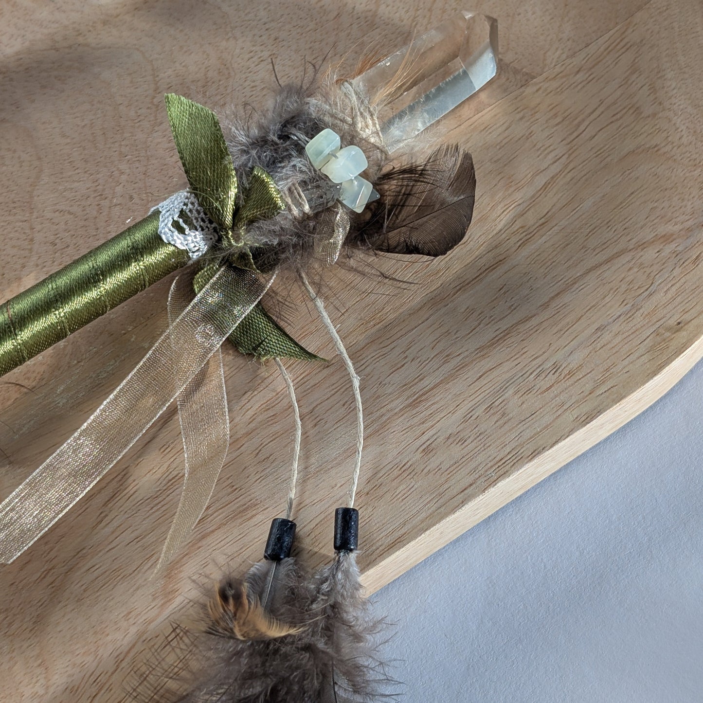 Handgefertigter Zauberstab mit einem goldgelben Citrin und schimmerndem Drusenquarz, verziert mit Federn und einem grünen Band, auf einer Holzplatte.