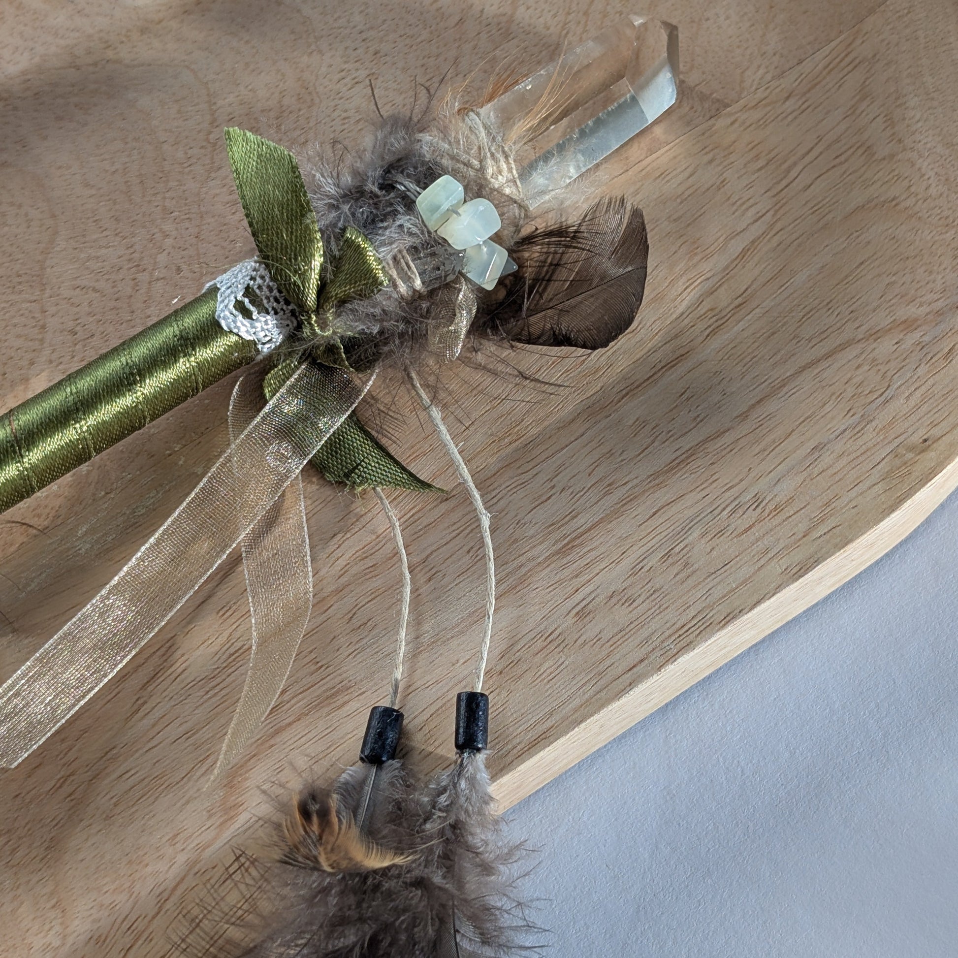 Handgefertigter Zauberstab mit einem goldgelben Citrin und schimmerndem Drusenquarz, verziert mit Federn und einem grünen Band, auf einer Holzplatte.