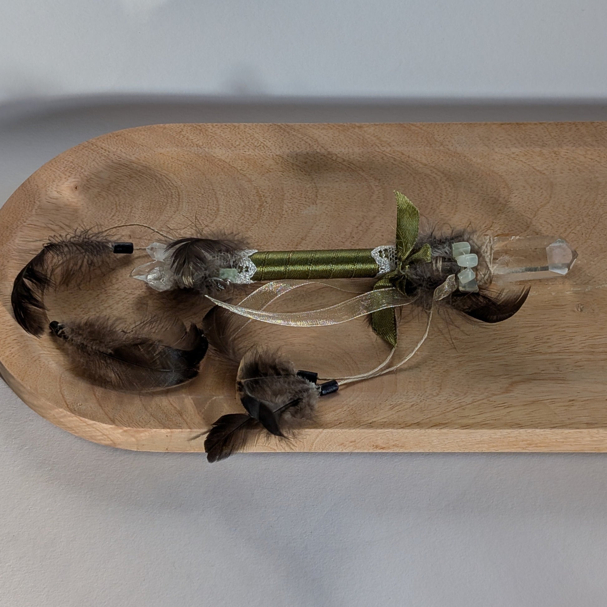 Handgefertigter Zauberstab mit einem goldgelben Citrin und schimmerndem Drusenquarz, verziert mit Federn und einem grünen Band, auf einer Holzplatte.