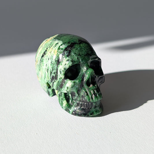 Kleiner Kristallschädel "Leonidar" (92g) aus grünem Zoisit mit schwarzen Einschlüssen.