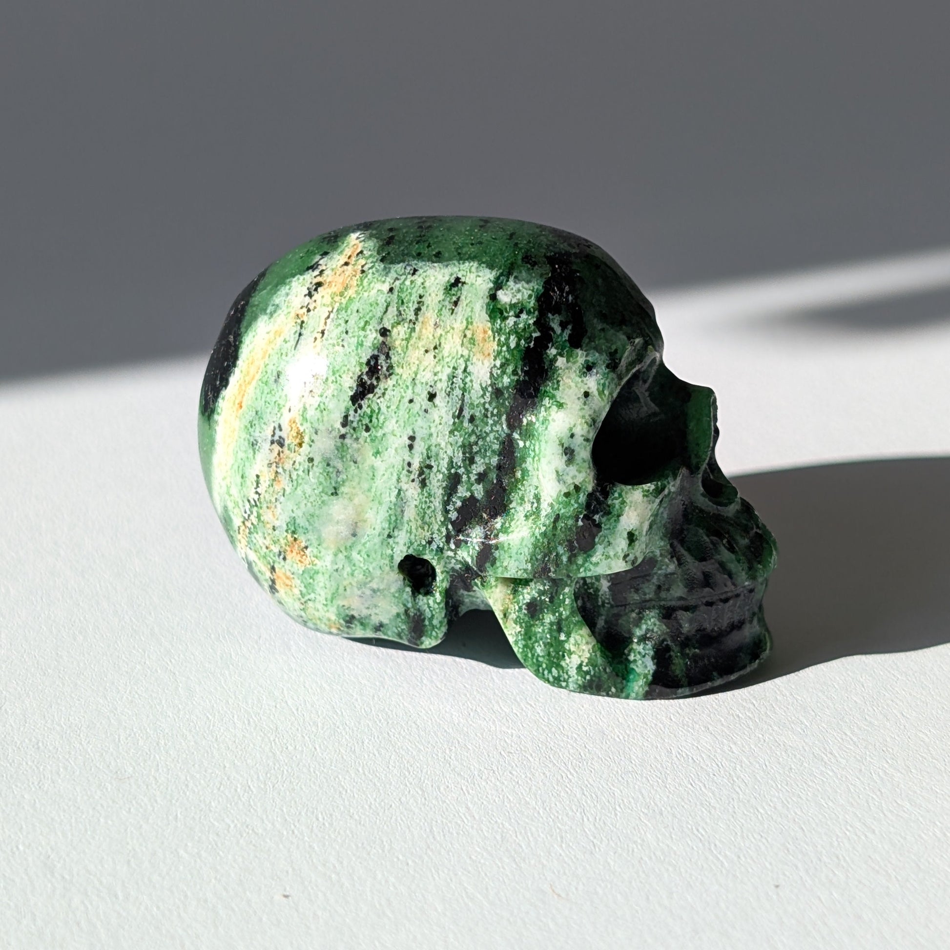 Kleiner Kristallschädel "Leonidar" (92g) aus grünem Zoisit mit schwarzen Einschlüssen.