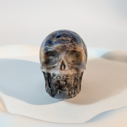 Massiver Kristallschädel Brok aus Sodalith mit zerrissener, tiefblauer, aschgrauer und rostig oranger Gesteinsmatrix.
