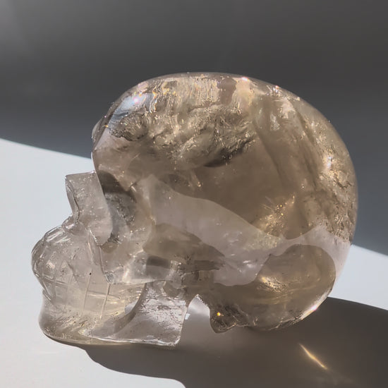 Ein 594g schwerer, klarer Rauchquarz-Schädel 'Der Wächter der Schatten' mit leuchtenden Regenbogen-Einschlüssen.